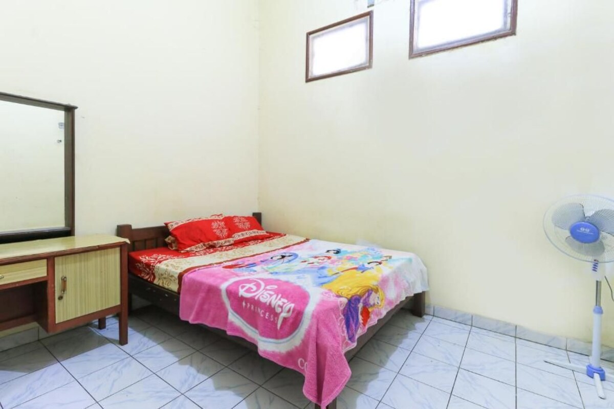 A bedroom with a bed, a desk, a mirror, and a standing fan.