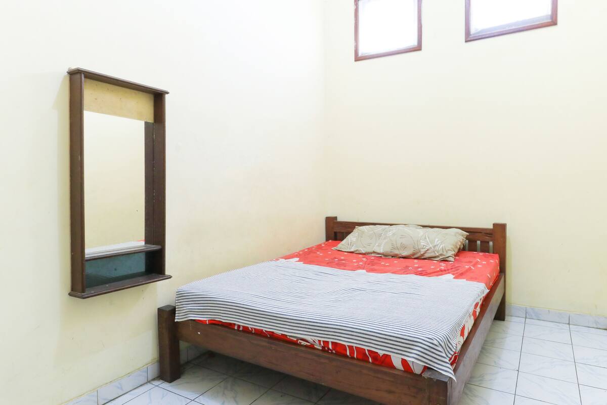 A small, neatly made bed with a red and white striped bedspread and a single pillow. A wooden headboard and a small shelf are attached to the wall.
