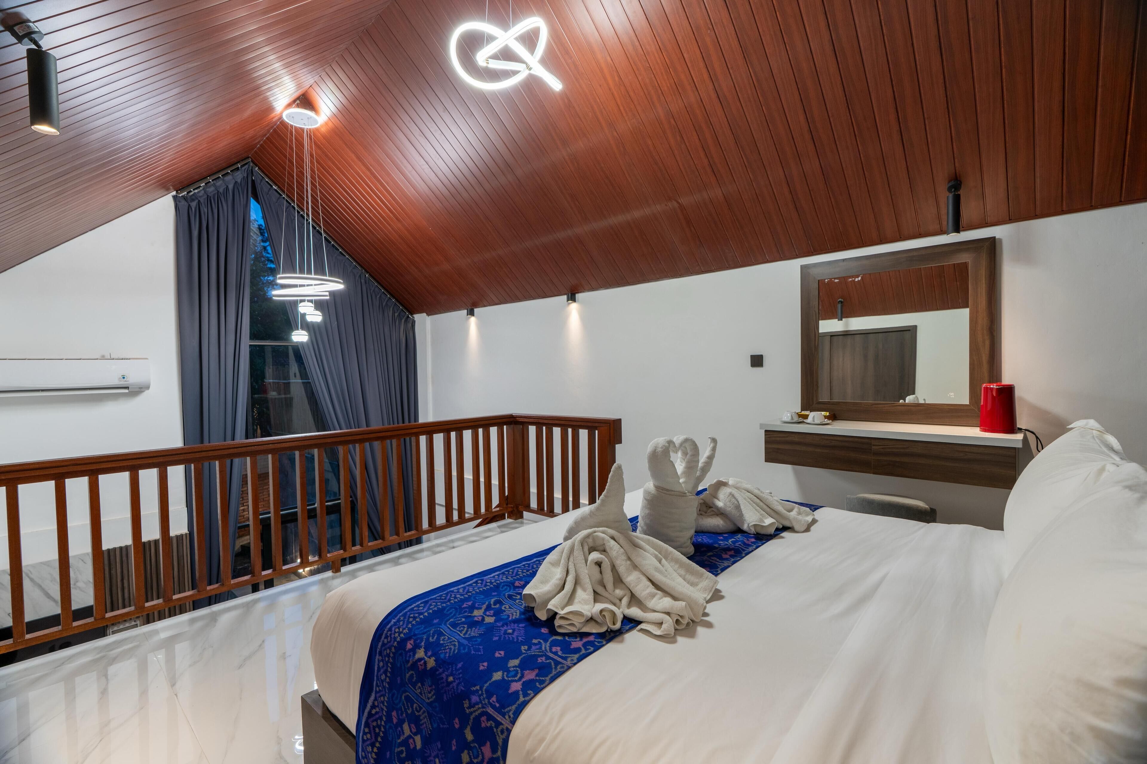 Une chambre d’hôtel avec un plafond en bois, un lit recouvert d’une couvre-lit à motifs bleus, une rambarde en bois, une table de chevet avec une tasse rouge et un miroir au-dessus du lit.
