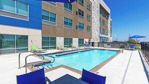 A swimming pool area with lounge chairs and umbrellas, surrounded by a building with multiple windows and a stone facade. - Holiday Inn Express & Suites Purcell by IHG (Purcell)