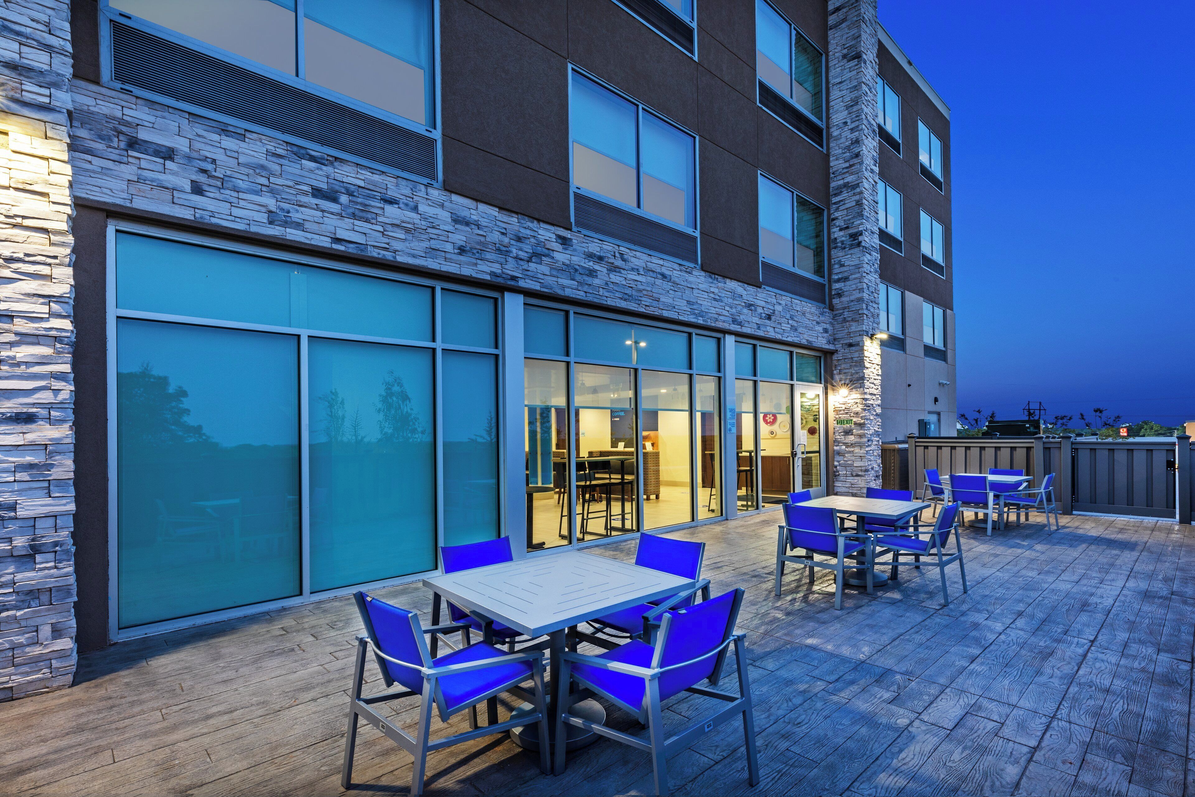 An outdoor seating area with tables and chairs, a stone facade, and large windows.
