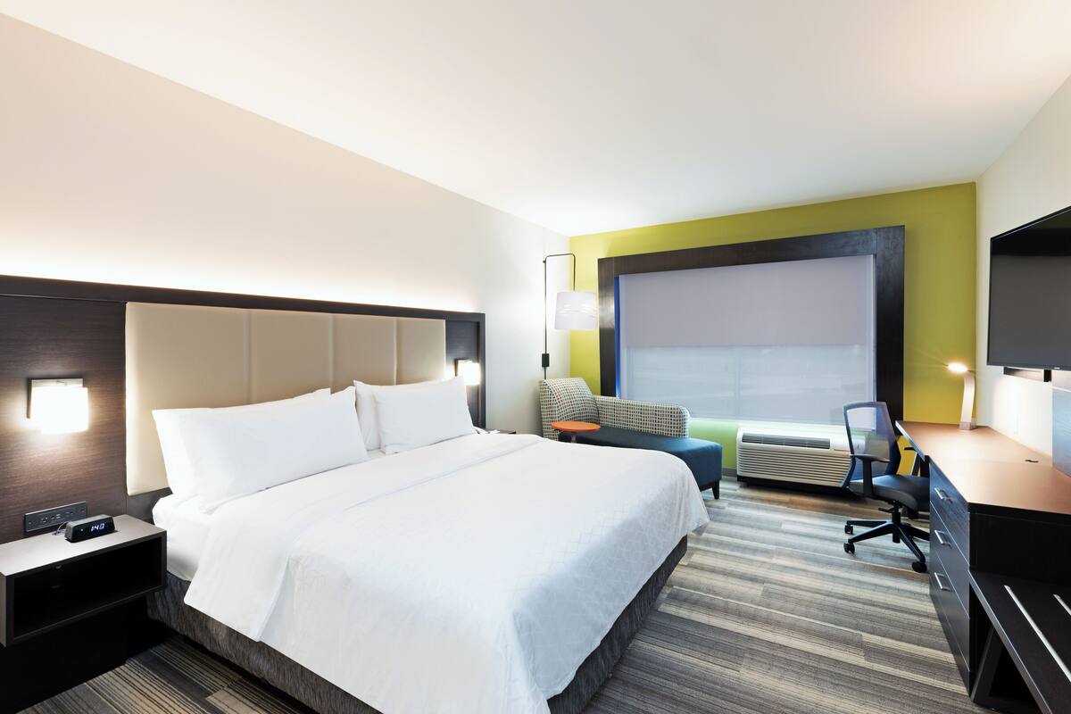 A hotel room with a large bed, a desk with a chair, a sofa, a TV, and a window with blinds.