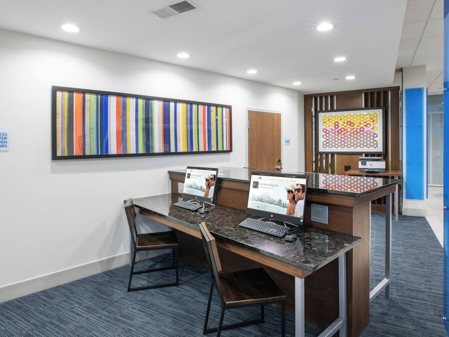 A reception desk with two computer monitors, a sign indicating business center hours, and a colorful abstract painting on the wall.