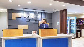 Un comptoir d’accueil d’hôtel avec un homme derrière, un écran numérique affichant le nom de l’hôtel et un distributeur automatique de collations.