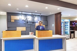 A hotel reception desk with a man standing behind it, a digital screen displaying the hotel's name, and a vending machine with snacks.