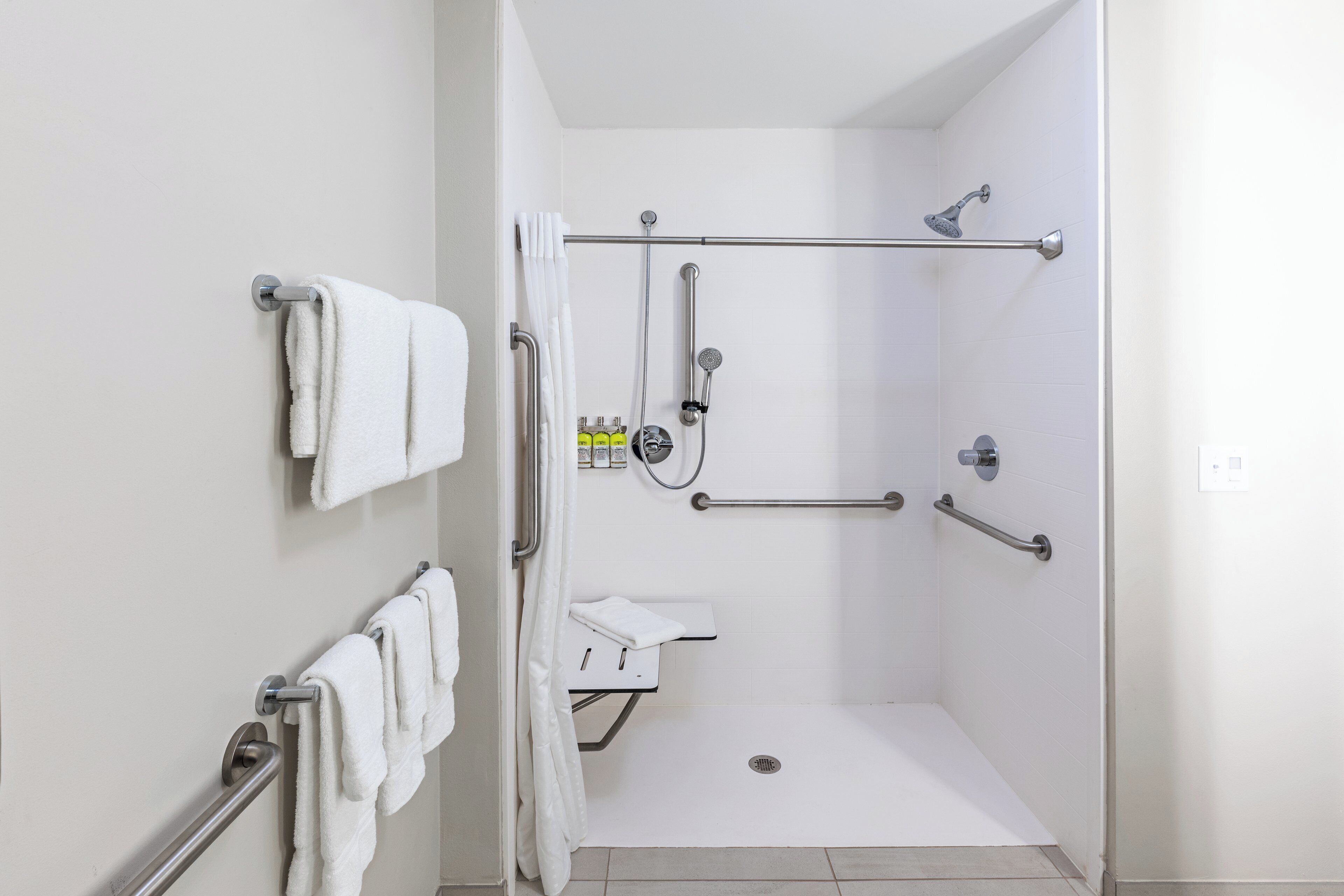 A wheelchair-accessible shower with grab bars, a white tiled floor, and a white towel rack.