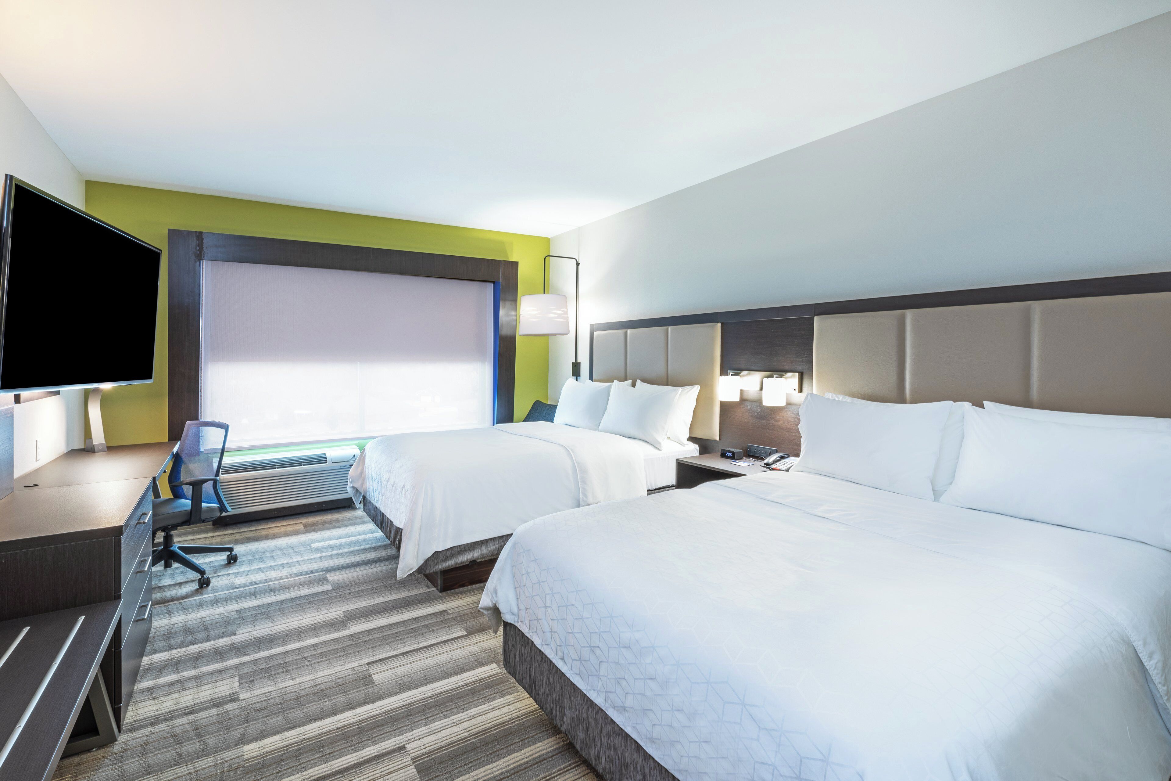 Ein Hotelzimmer mit zwei Betten, einem Schreibtisch, einem Stuhl, einem Flachbildfernseher und einem großen Fenster.