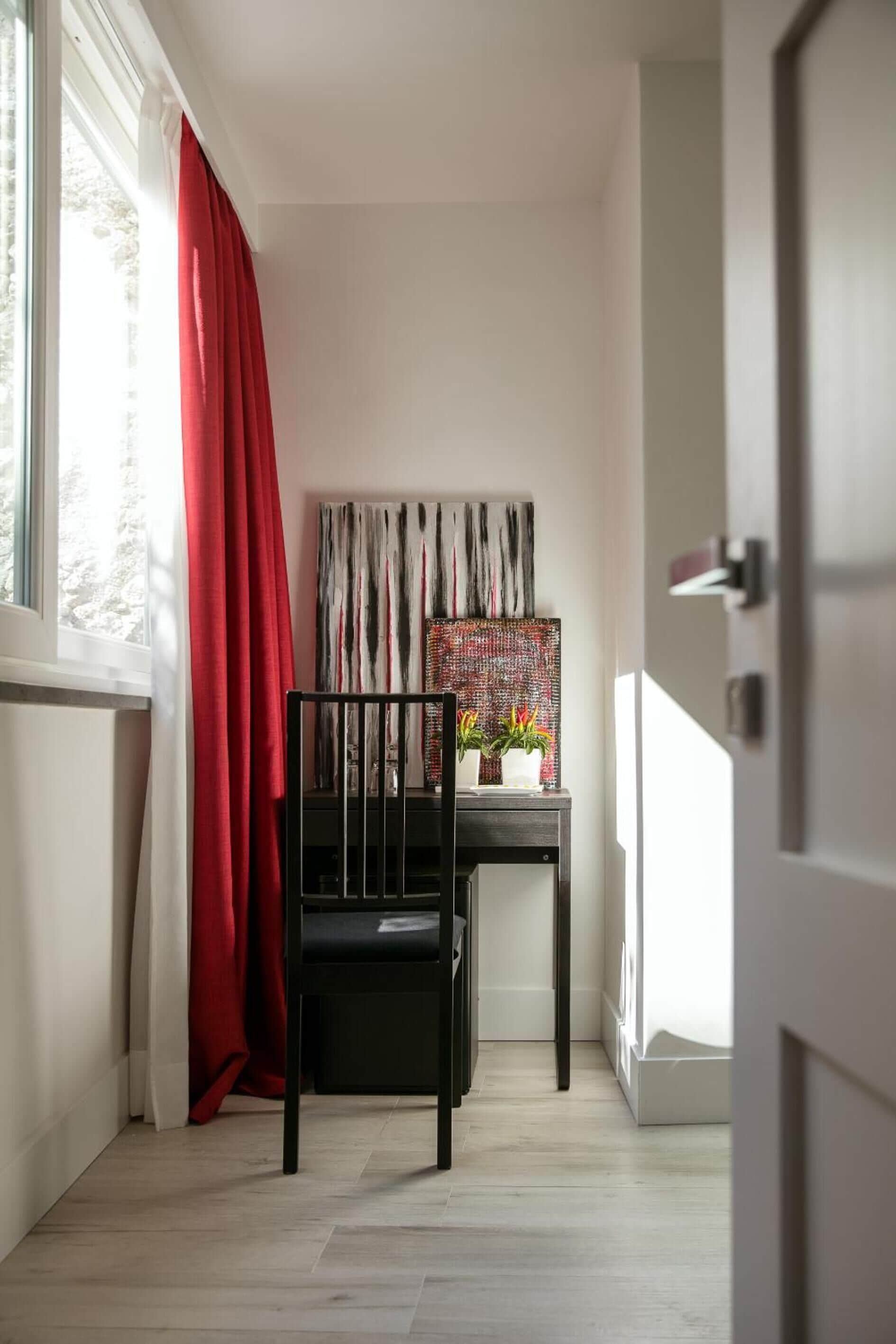 Een kamer met een zwarte stoel, een tafel met een vaas en planten, een rood gordijn en een schilderij aan de muur.