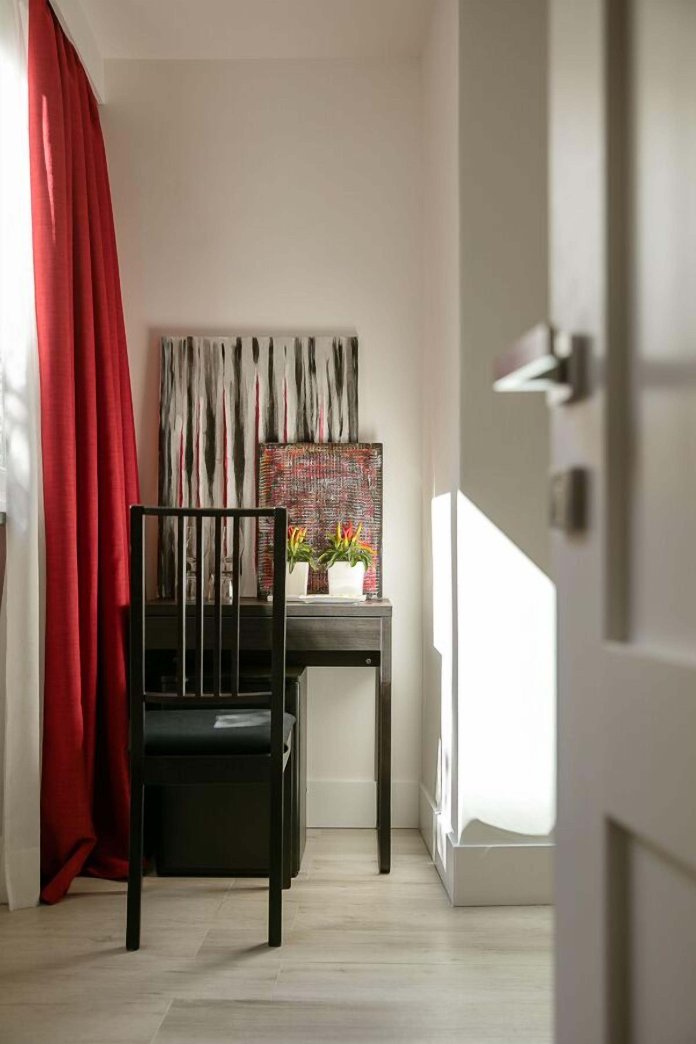 Una habitación con una silla negra, un escritorio con un cuadro abstracto colorido y una cortina roja.