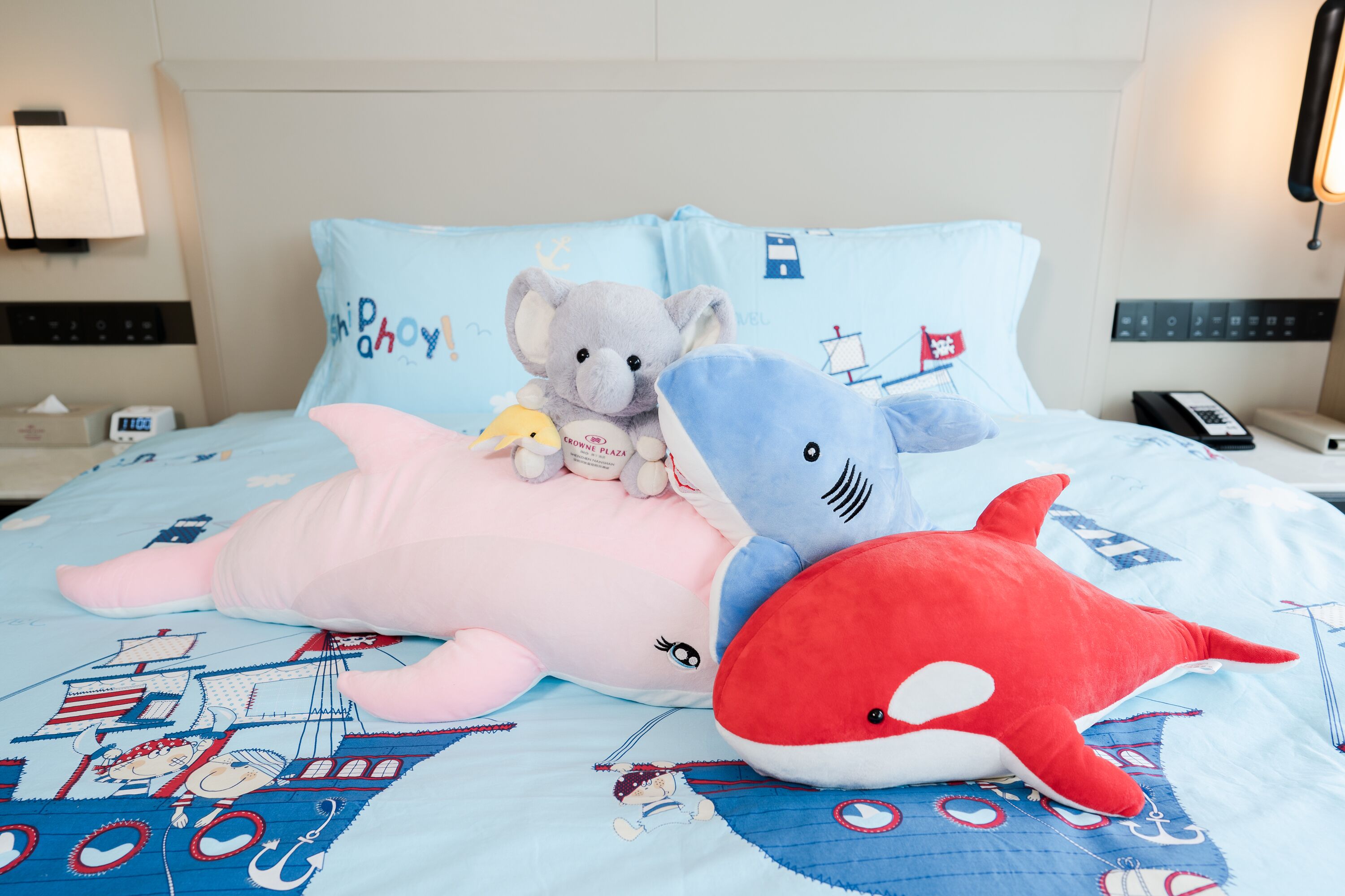 A bed with a blue quilt featuring maritime designs, a pink stuffed animal, a grey stuffed elephant, and a red stuffed shark.