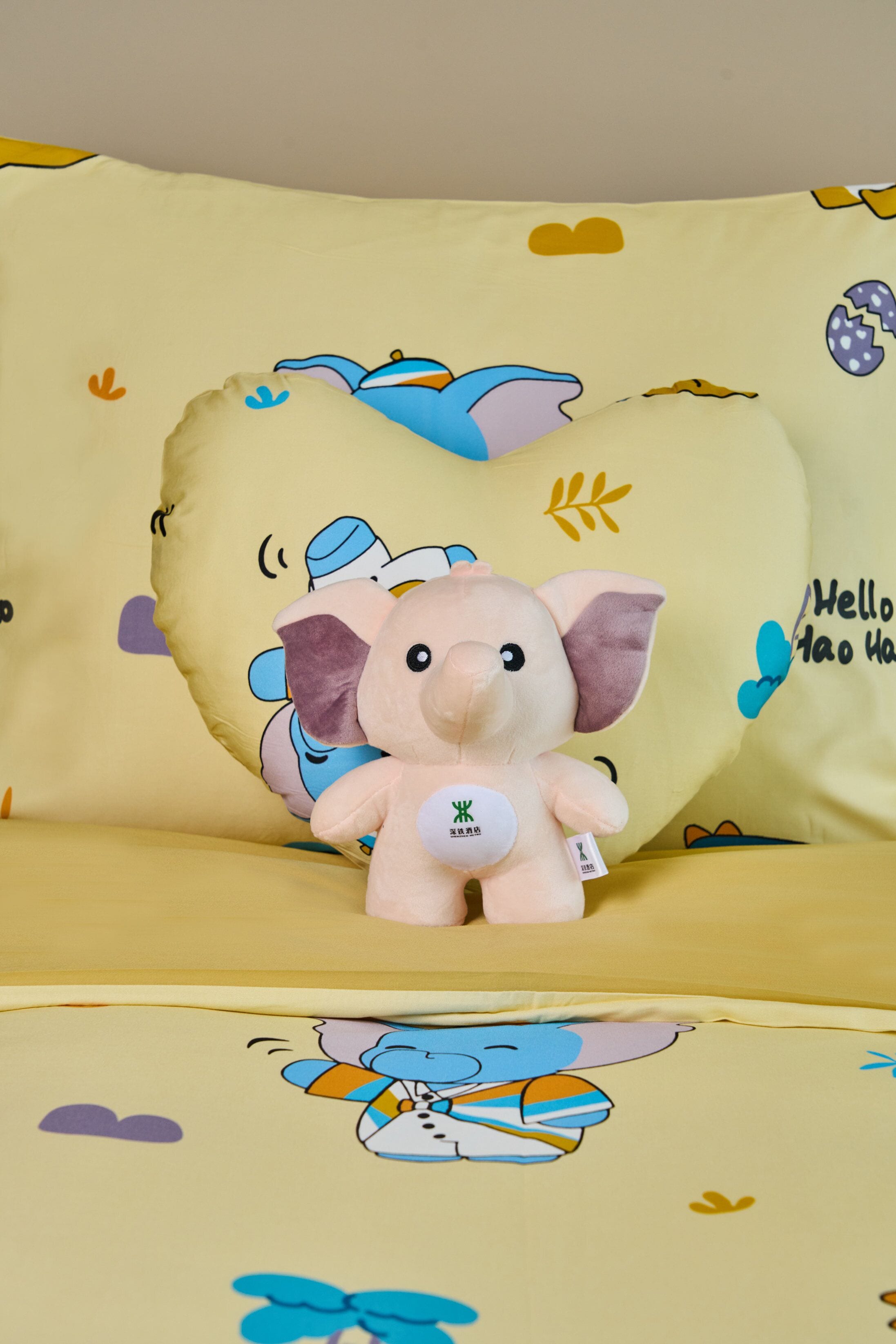 Un elefante de peluche sobre un almohadón amarillo con un diseño de corazón e ilustración de un pájaro azul.