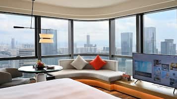 A modern hotel room with a curved bed, a seating area with cushions, a television, and a large window offering a city view.