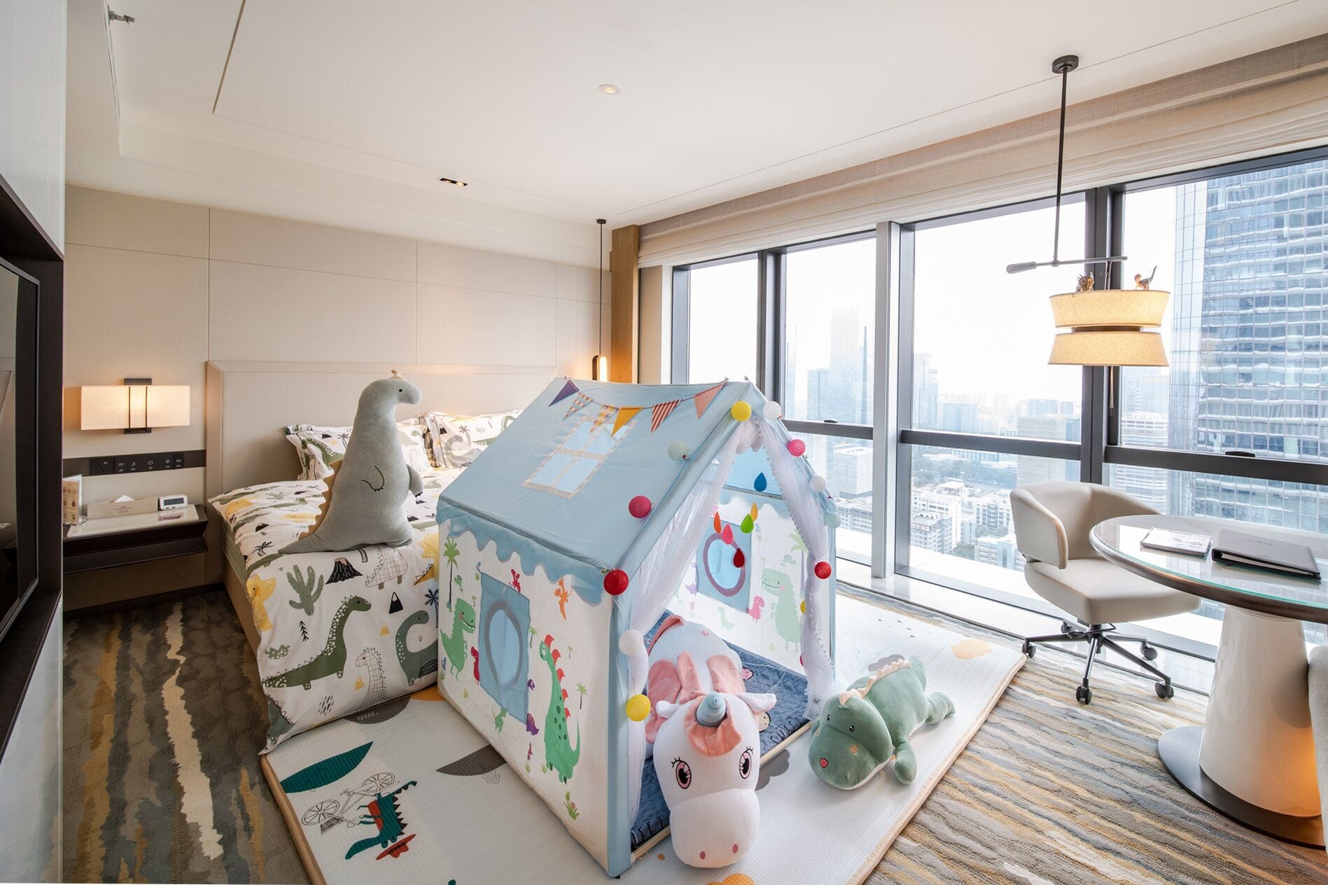 Une chambre d’hôtel avec une grande fenêtre, un lit avec une couverture à motifs de dinosaures, une tente de jeu et un bureau avec une chaise.