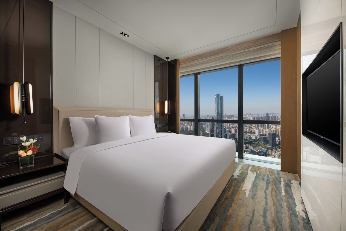 A modern hotel room with a large bed, a TV, a nightstand, and a city view.