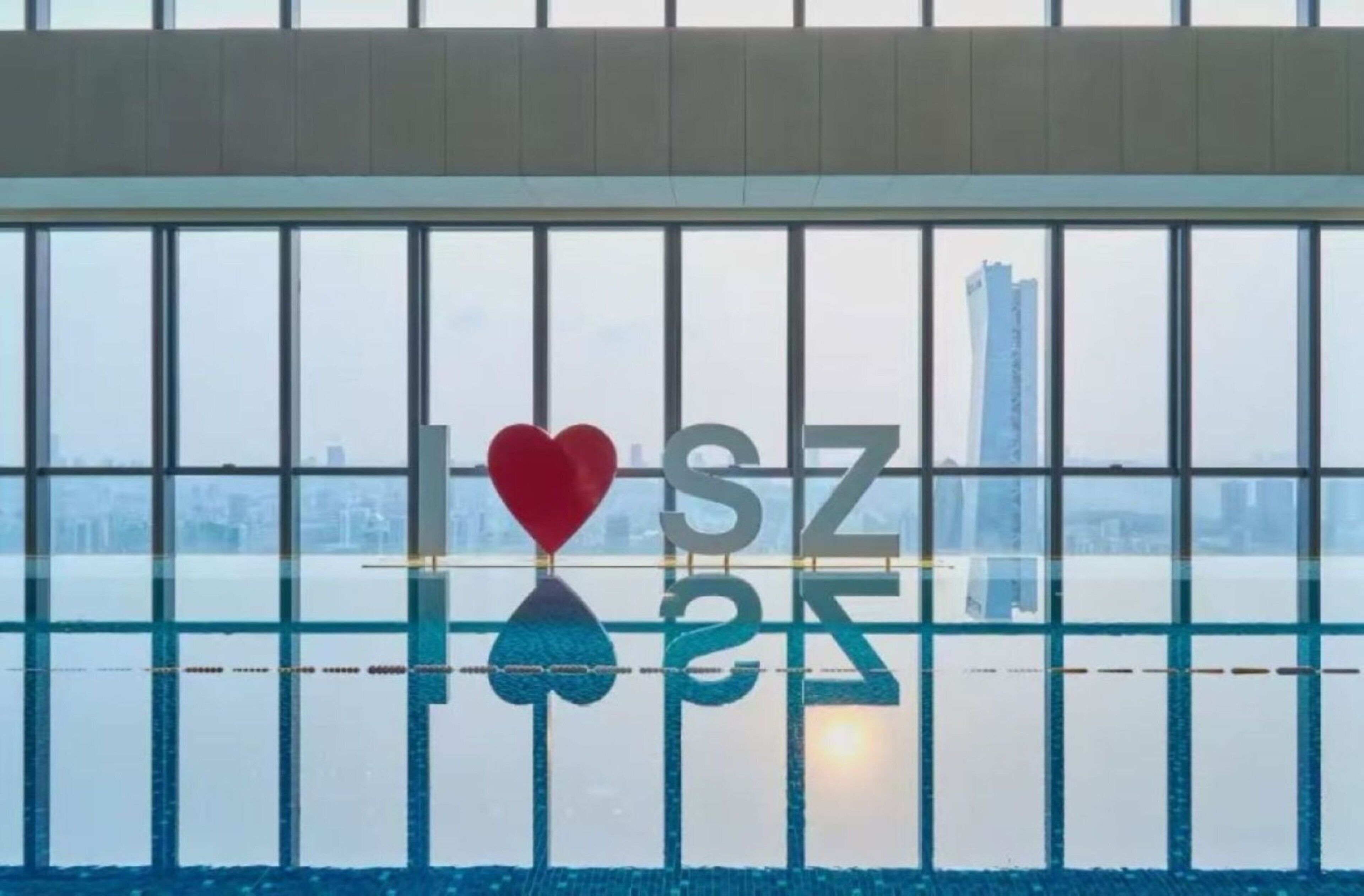 A view through a window with a reflection of a cityscape and a building with the letters 'IHSZ' and a heart symbol.