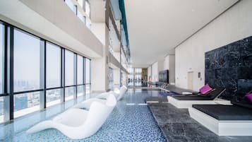 A modern indoor swimming pool with a view of the city, lounge areas with seating, and a large black marble wall.