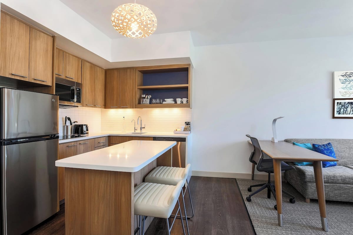 Une cuisine moderne avec des meubles en bois, un réfrigérateur en acier inoxydable, un four à micro-ondes et un évier. On y trouve un petit îlot avec des tabourets et une table avec des chaises. Un canapé et un fauteuil sont visibles au fond.