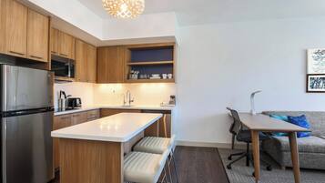 Een moderne keuken met houten kastjes, een roestvrijstalen koelkast, een magnetron en een spoelbak. Er is een klein eiland met krukken en een tafel met stoelen. Een bank en een stoel zijn zichtbaar op de achtergrond.