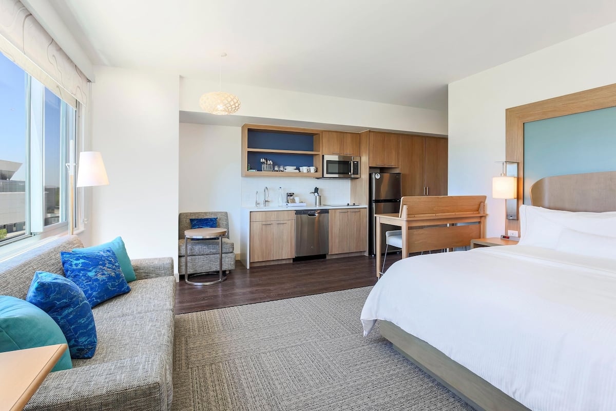Une chambre d’hôtel moderne avec un grand lit, un canapé avec des coussins bleus, un petit coin cuisine équipé d’un micro-ondes et d’un réfrigérateur, et une vue sur la ville par la fenêtre.