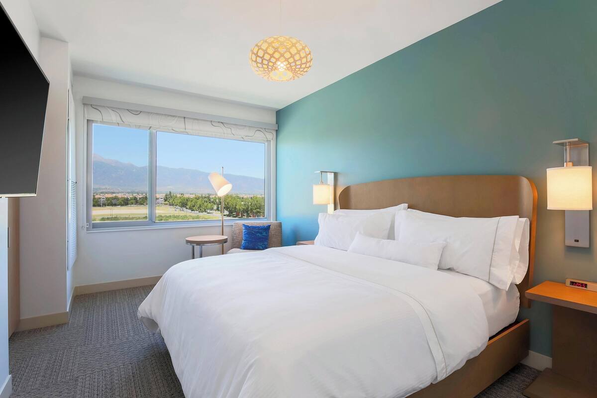 Une chambre d’hôtel avec un grand lit, une table de chevet avec une lampe, une petite table avec une chaise, et une vue sur les montagnes par la fenêtre.