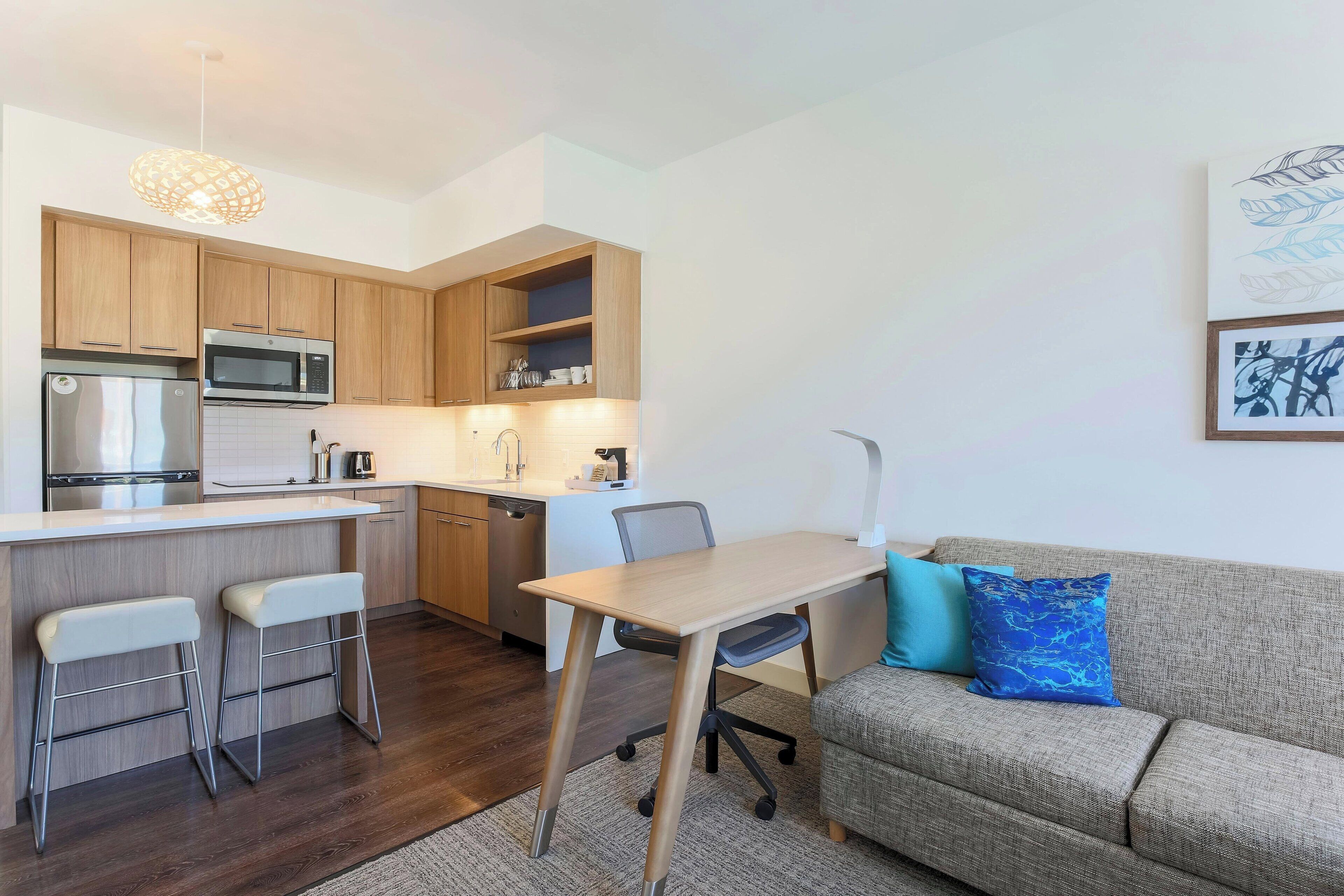 Eine moderne Küche mit Holzausstattung, einer Mikrowelle und einem Spülbecken. Ein Essbereich mit Tisch und zwei Barhocker. Ein Sofa mit blauem Kissen lehnt an der Wand.