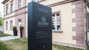 A black signboard with the text "HPP Projekt Bauträger GmbH" and "ALTE SCHULE BOARDING HOUSE" along with contact information.