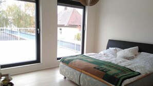 A bedroom with a large window, a bed with a patterned bedspread, and a view of a balcony and buildings outside.