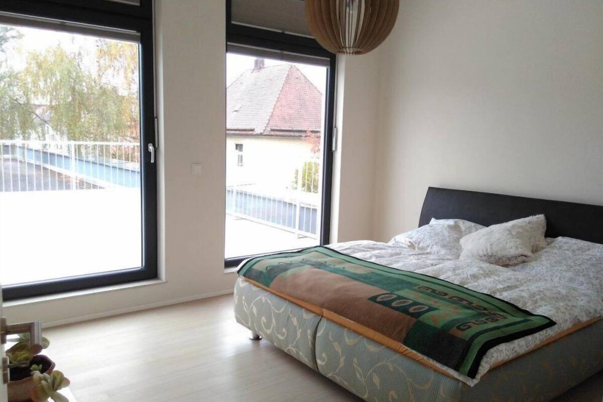 Een slaapkamer met een groot raam, een bed met een beddeken met een patroon, en uitzicht op een balkon en gebouwen.