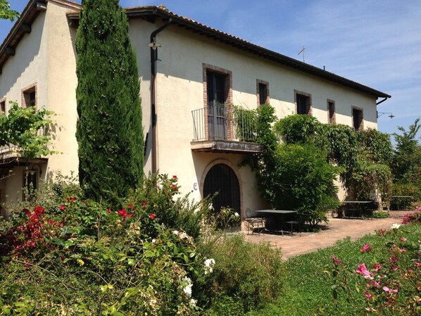 Stroll through the charming grounds, surrounded by lush greenery and vibrant flowers, to the entrance of this two-story building with a balcony and arched doorway.