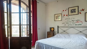 Une chambre à coucher avec un lit, une porte en bois et une fenêtre ornée de rideaux rouges. Des tableaux encadrés sont accrochés au mur, et une petite table avec une lampe s'y trouve.