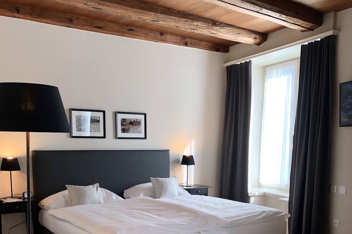 A bedroom with a large bed, a bedside table with a lamp, a chair, and a window with curtains.