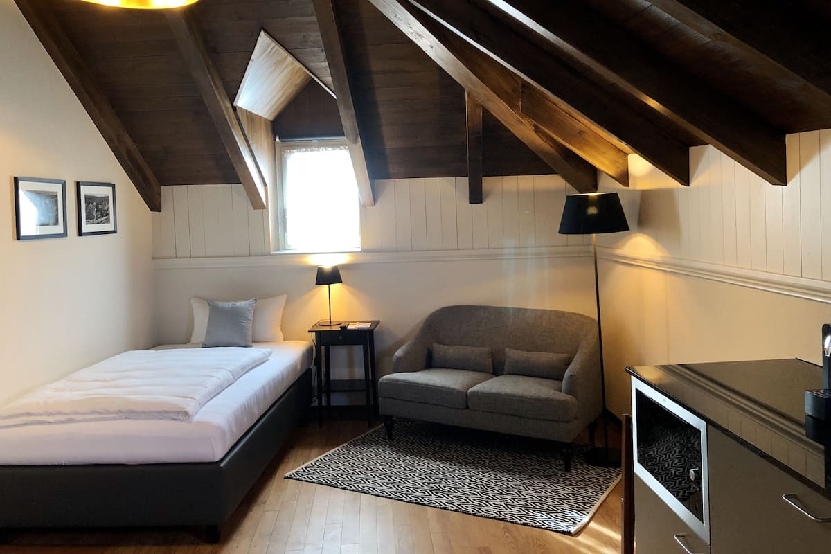 A bedroom with a bed, a grey sofa, a small table with a lamp, and a TV.