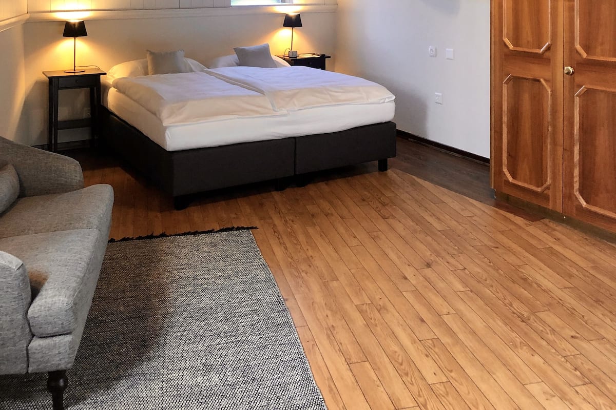 A bedroom with a sloped ceiling, wooden beams, a bed with white bedding, two bedside lamps, a TV mounted on the wall, and a wardrobe.