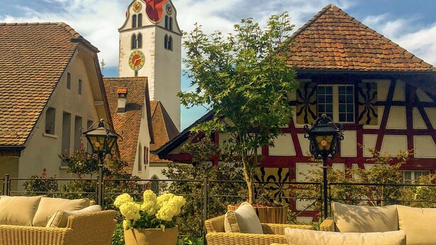 A terrace with wicker furniture and a large planter, set against a backdrop of traditional buildings and a church tower. - Marco Polo Business Apartments Sonne - Bremgarten (Bremgarten)
