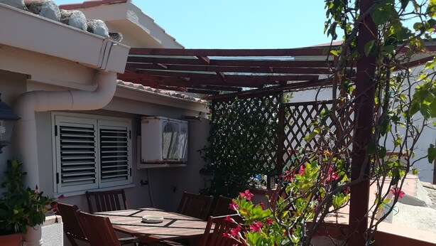 Un patio al aire libre con una mesa y sillas, rodeado por una pérgola y plantas en macetas.