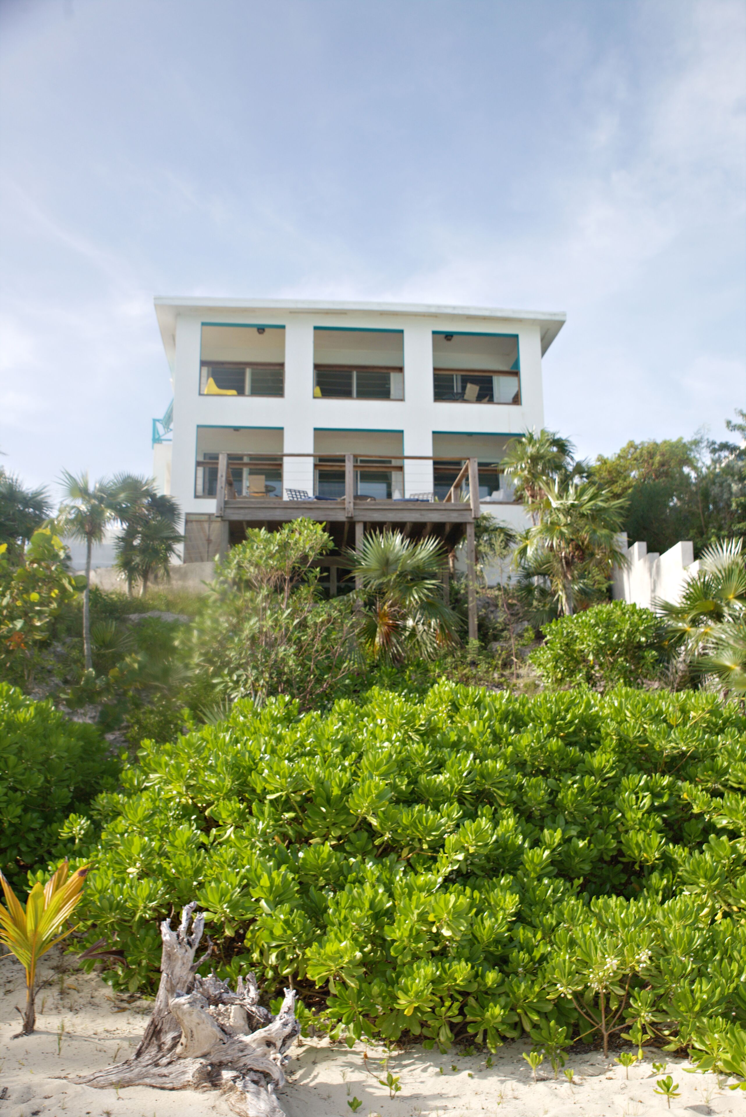 A multi-story building with a flat roof and a balcony, surrounded by dense green vegetation and a sandy foreground.