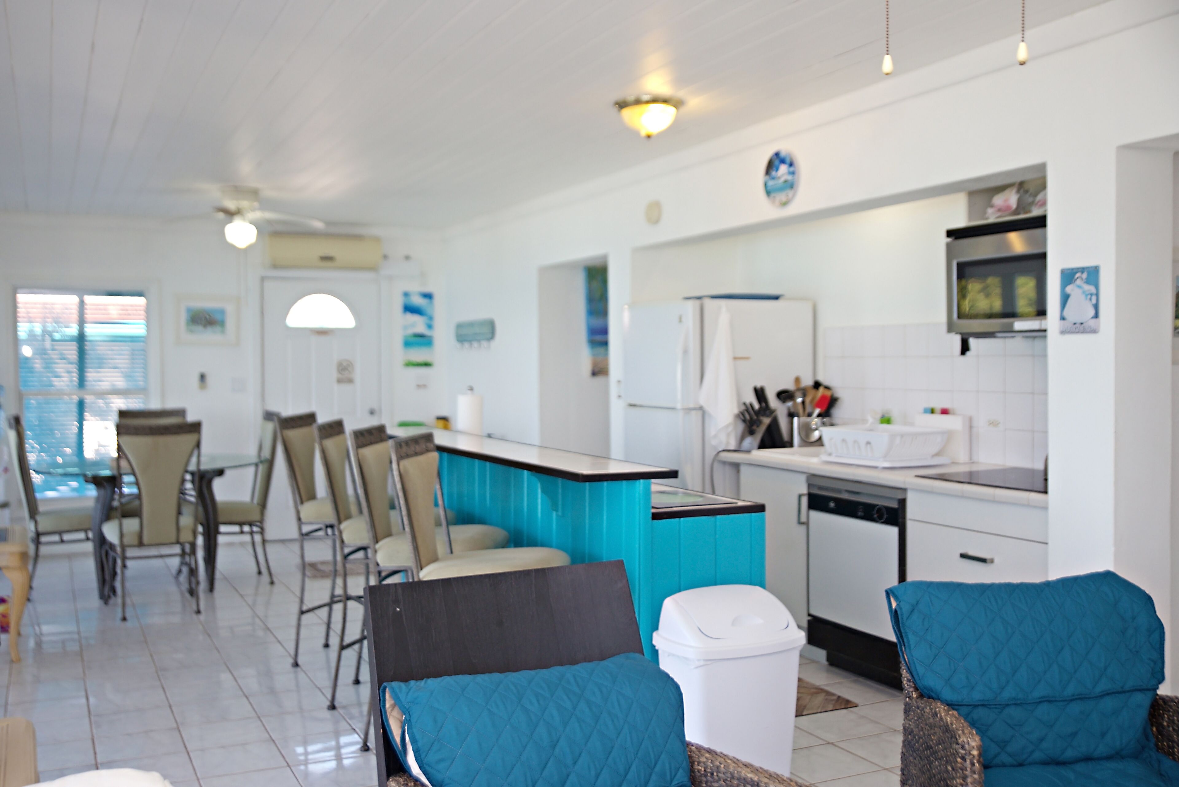 A modern kitchen with white cabinets, a blue countertop, and a dishwasher. There are two blue chairs and a white trash bin.
