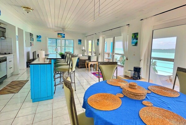 Un amplio salón con una mesa azul, individuales de mimbre y vista al océano.