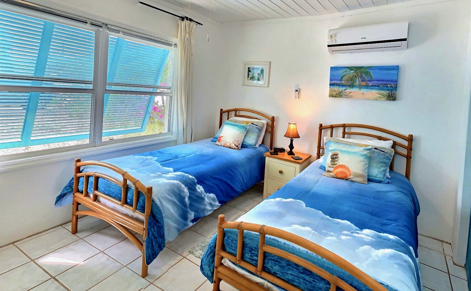 Un dormitorio con dos camas de madera, cada una con una colcha azul con diseño de nubes. Hay una ventana con persianas, un aire acondicionado y una mesita de noche con lámpara.