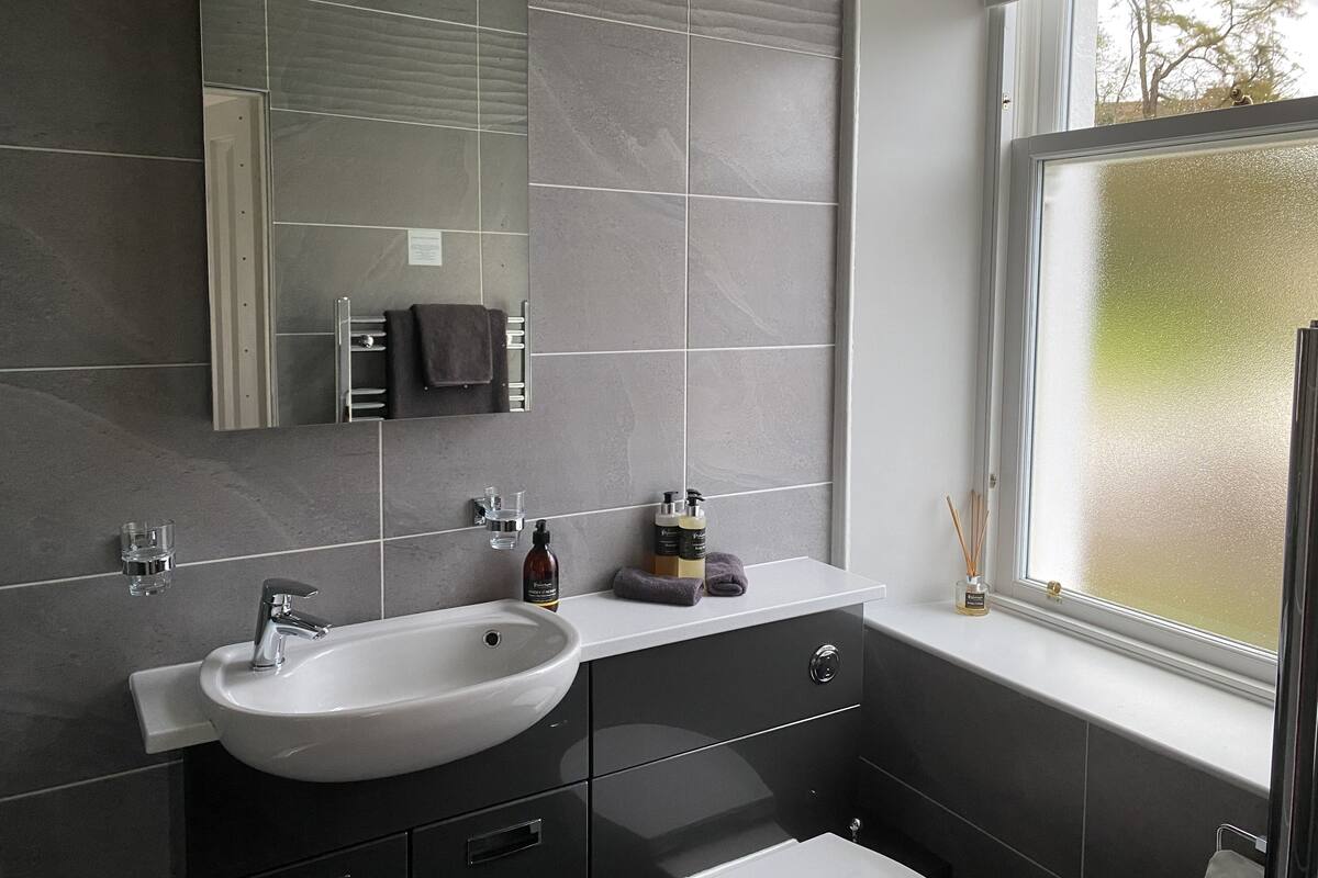 Een moderne badkamer met een grote spiegel, een witte wastafel, donkere kastjes en een raam met uitzicht op bomen.
