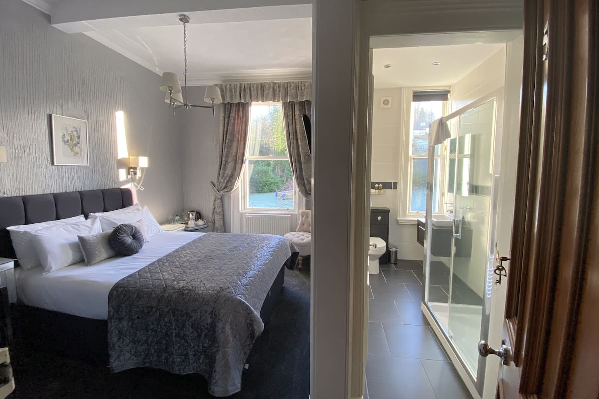 Een slaapkamer met een groot bed, een nachtkastje met een lamp, een raam met gordijnen, en uitzicht op een badkamer.