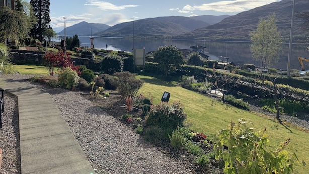 Een schilderachtig uitzicht op een meer, met bergen op de achtergrond, een verzorgde tuin en een heldere lucht.