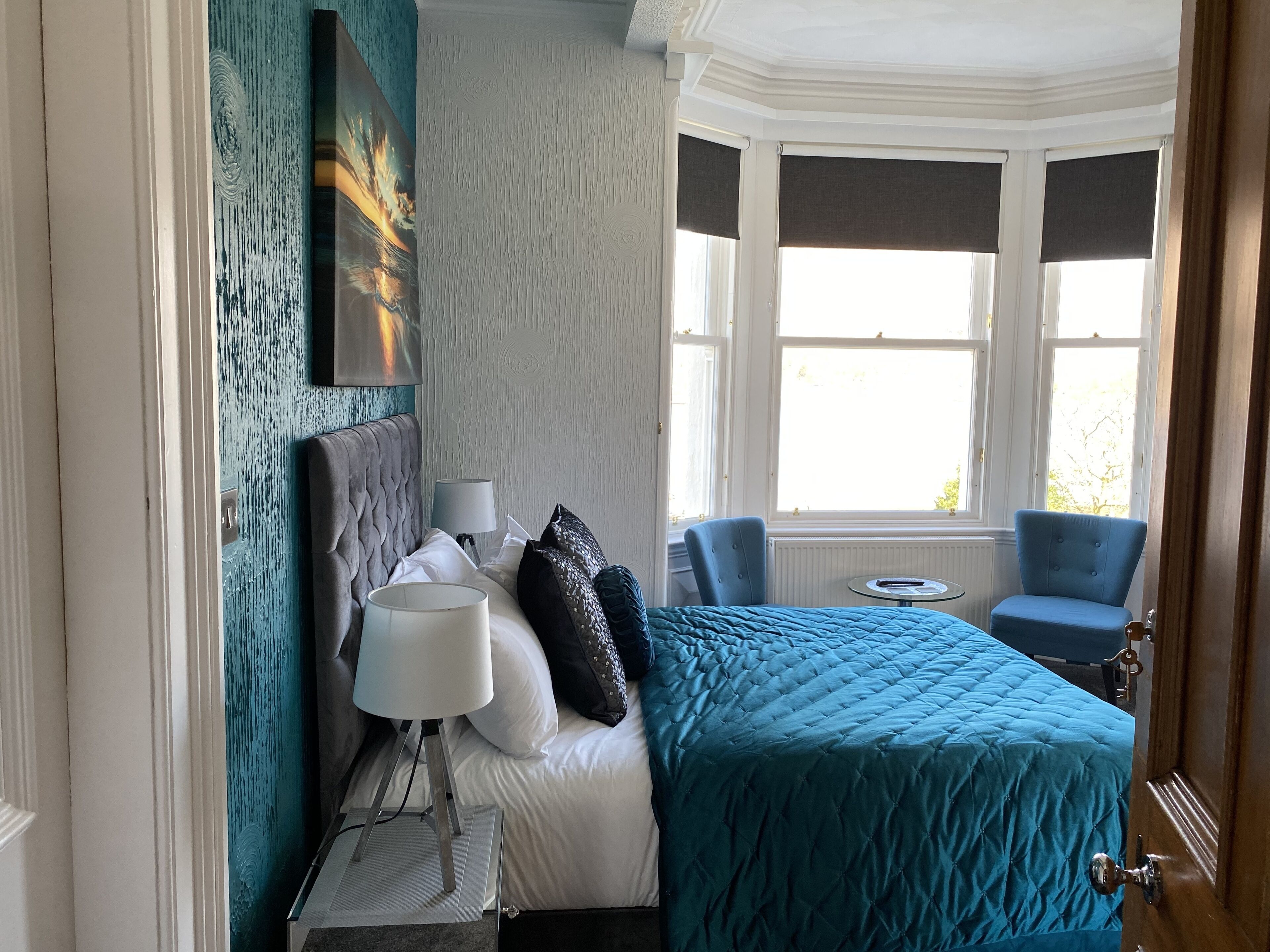 A hotel room with a large bed, a chair, a small table, and a bay window with curtains.
