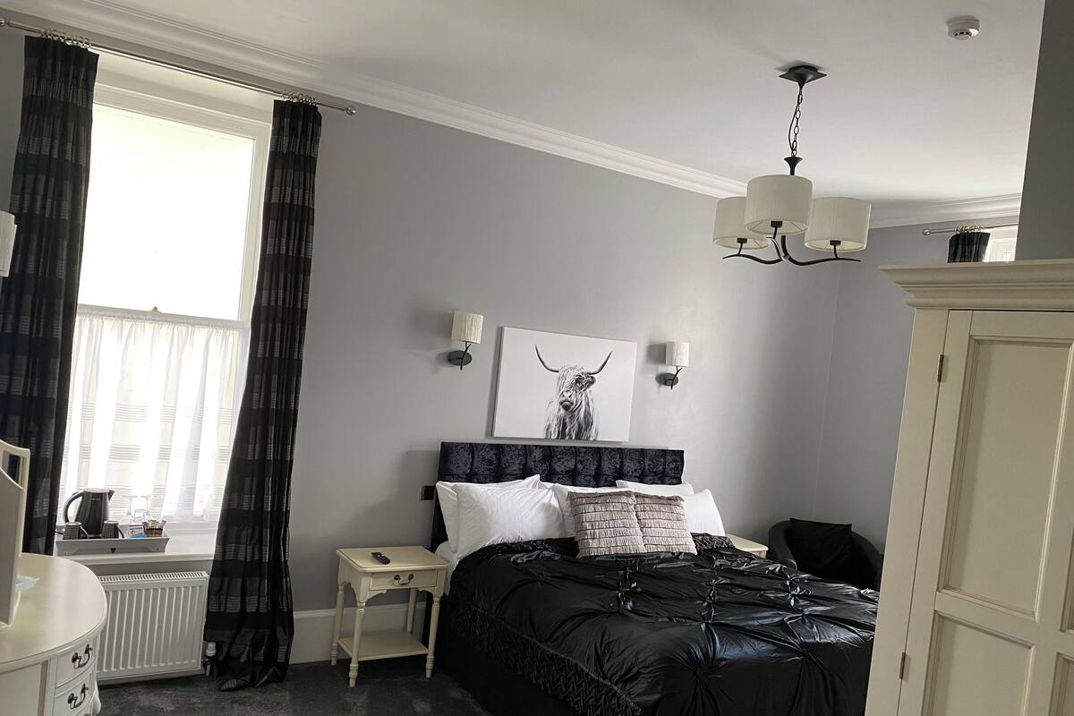 Een slaapkamer met een bed, nachtkastjes, een radiator, een raam met gordijnen, een wandlamp en een plafondlamp.
