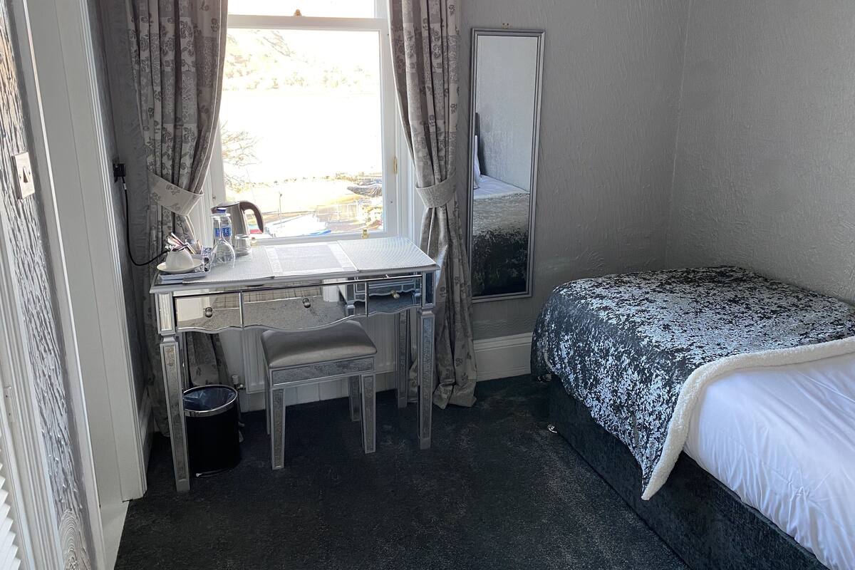 Een kleine, compacte kamer met een bureau en stoel, een bed met een bedovertrek met dessin, en een grote spiegel.