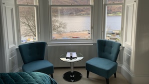 Een kamer met twee blauwe fauteuils, een tafeltje en uitzicht op een meer en bergen door grote ramen.