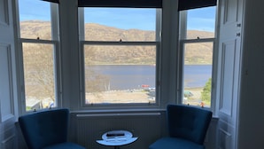 Een kamer met twee blauwe stoelen, een klein rond tafeltje en uitzicht op een meer en bergen door grote ramen.