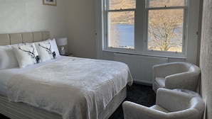 Een slaapkamer met een groot bed, twee stoelen, een nachtkastje en een raam met uitzicht op een meer en heuvels.
