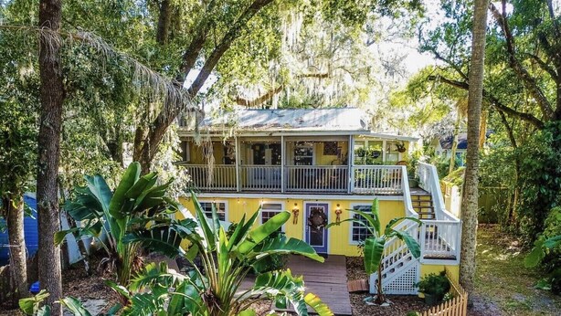 A two-story yellow house with a porch, surrounded by lush greenery and trees.