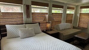 A room with a large bed, a sofa, a nightstand with a lamp, and a window with a view of trees.